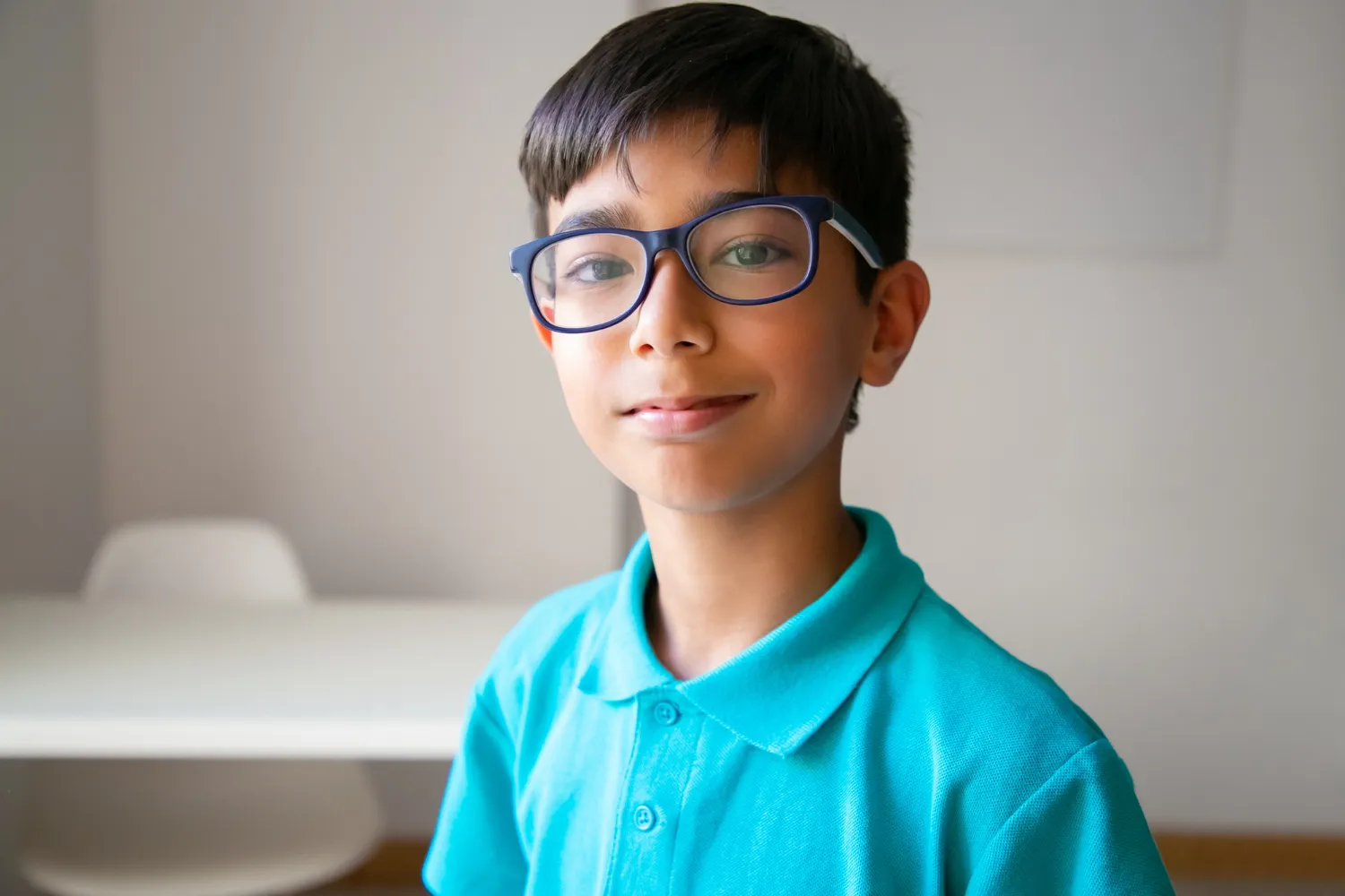 Smiling young boy wearing dark-rimmed glasses and a blue polo shirt, standing indoors.