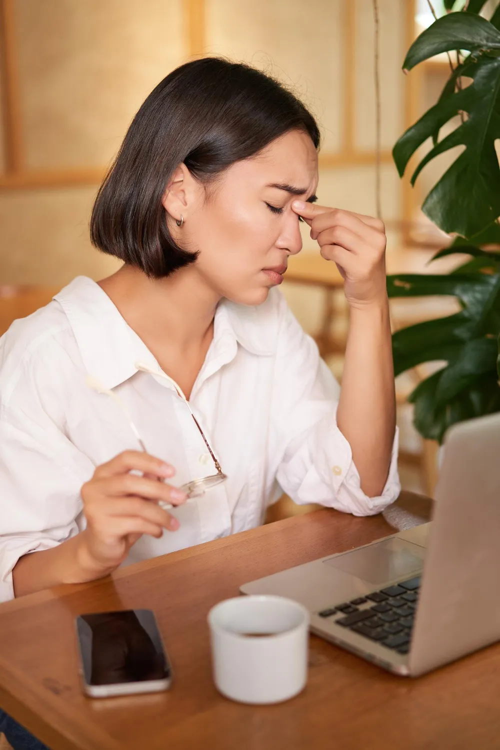 Stressed woman with short dark hair, wearing a white shirt, rubs her temples with eyes closed while holding glasses. A laptop is in front of her.