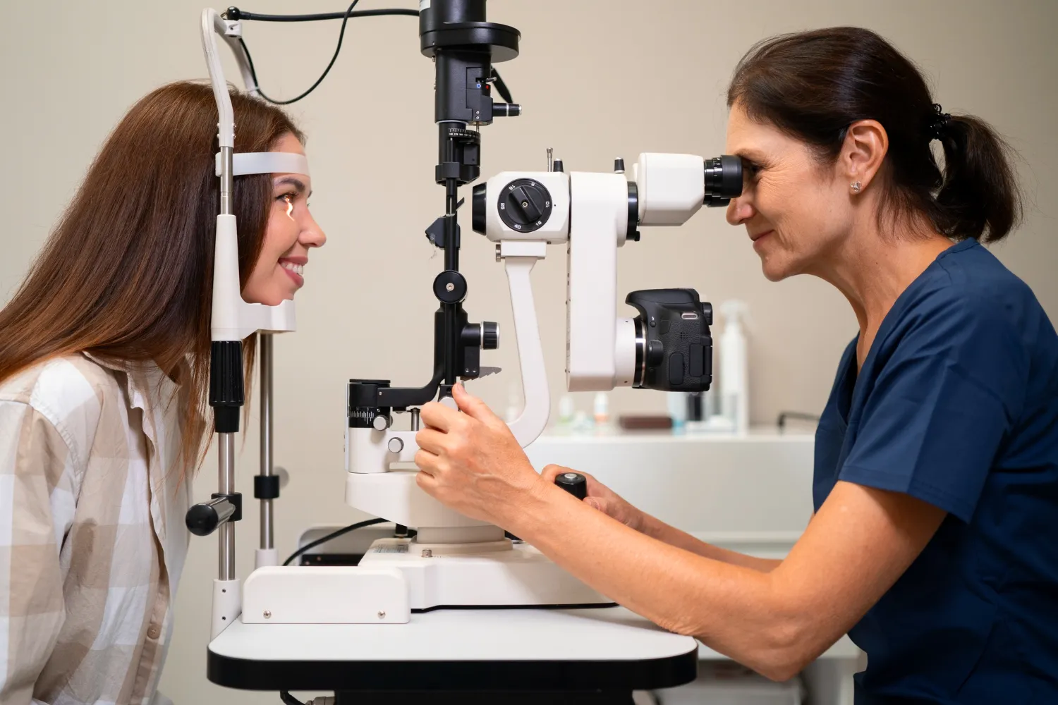 Optometrist examining a smiling patient's eye with a slit lamp microscope during a routine eye exam.