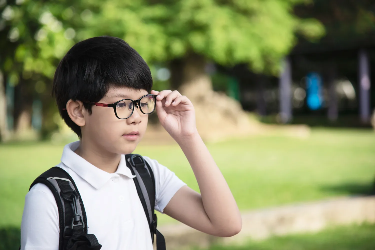 Young boy wearing a white polo shirt, backpack, and glasses, adjusting his spectacles outdoors in a park.