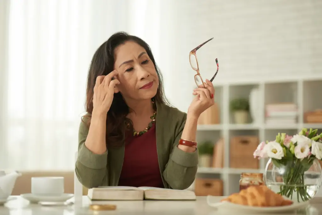 Middle-aged Asian woman pauses while reading a book, holding her eyeglasses and touching her temple in thought.
