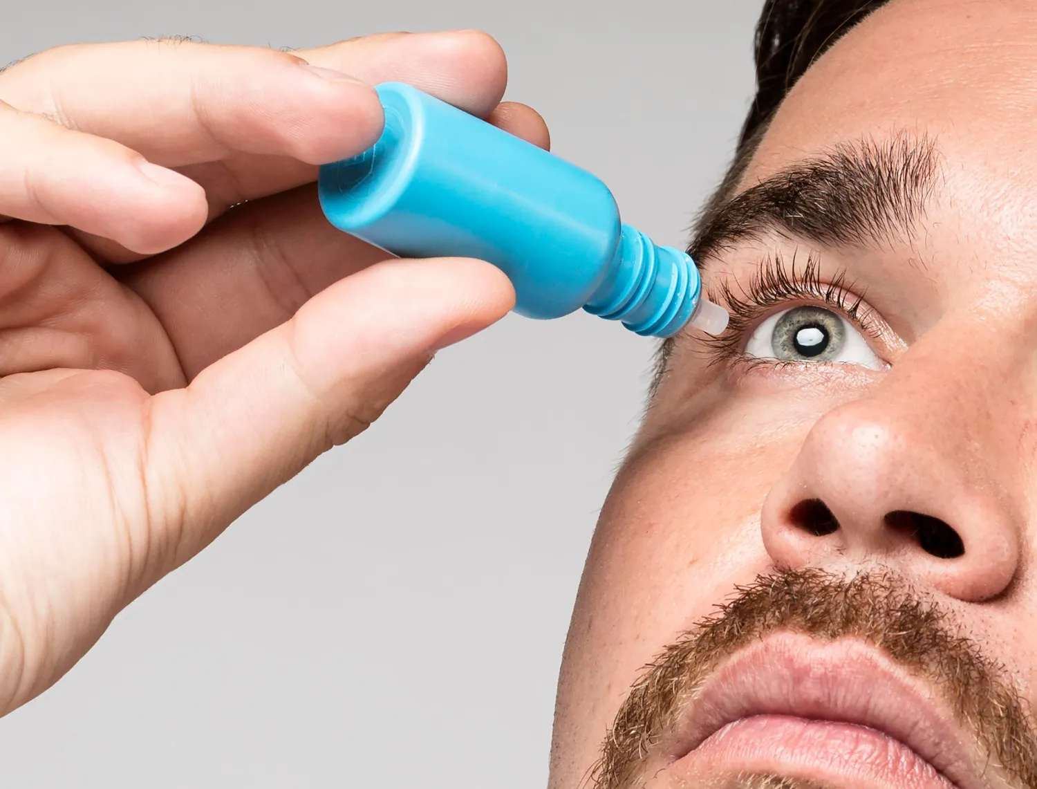 Close-up of a man applying eye drops to his open right eye with a blue bottle.
