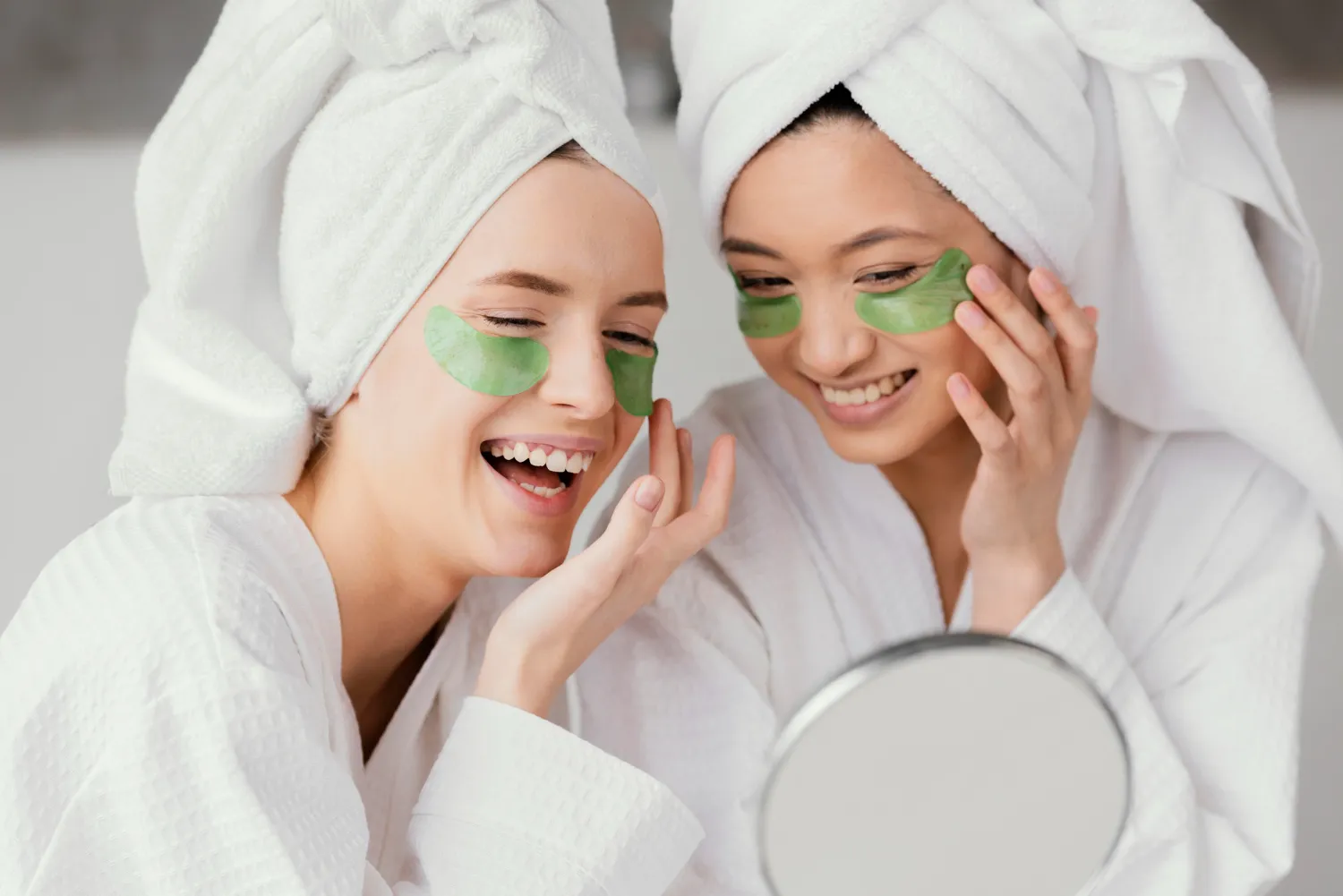 Two diverse women in white bathrobes and hair towels with green eye patches, laughing while looking in a mirror during a skincare routine.