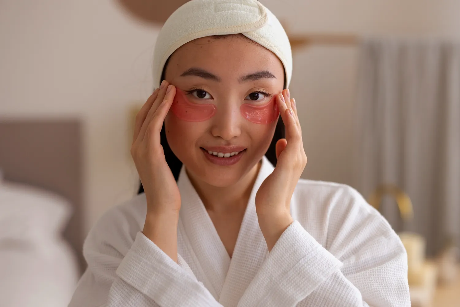 Smiling woman in a white bathrobe and headband, applying pink under-eye patches to her face.