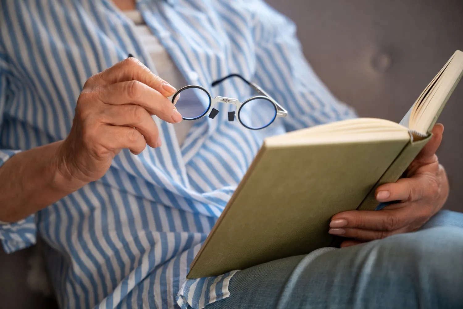 Close-up of a person holding round spectacles while reading an open book with a green cover.