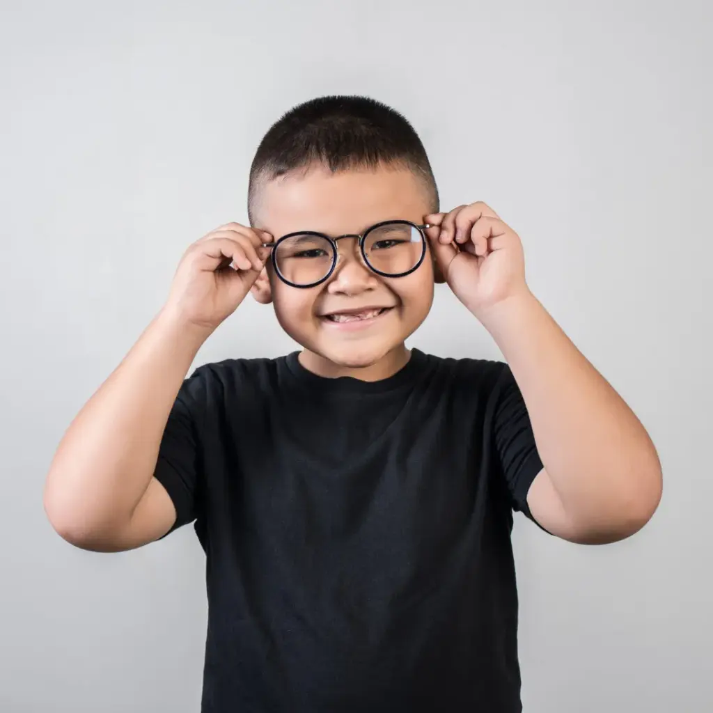 Smiling young boy wearing a black shirt, adjusting round glasses with both hands against a grey background.