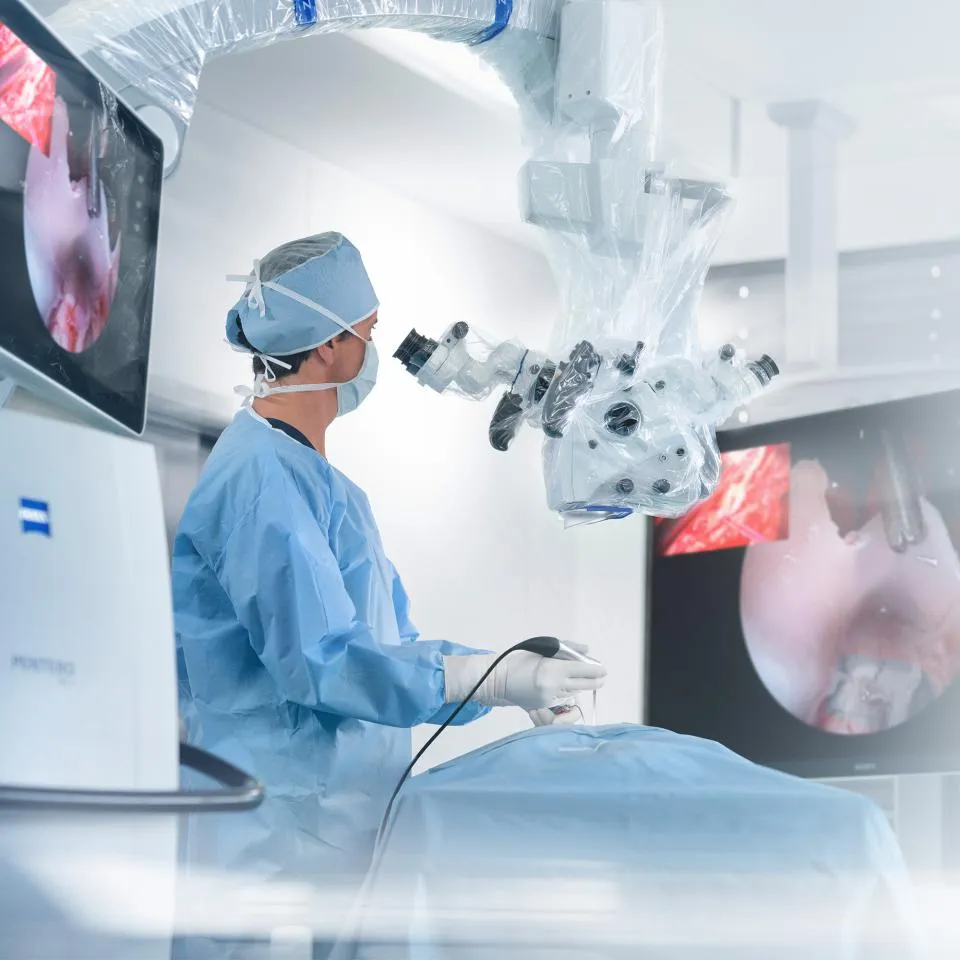 Surgeon in blue scrubs operates with a robotic surgical microscope, monitoring endoscopic views on screens.