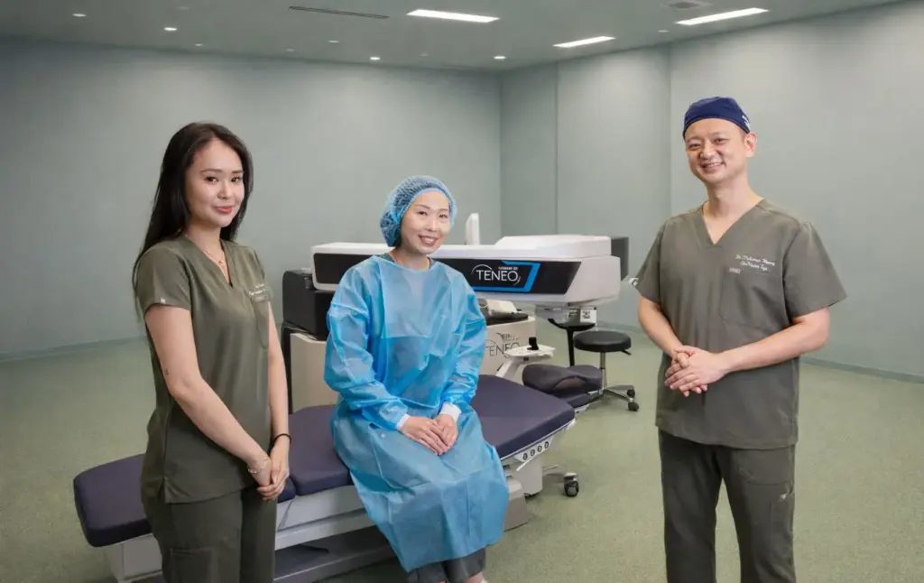 Smiling patient in a blue gown sits by a large eye surgery machine, flanked by two medical staff in green scrubs, in a modern clinic room.