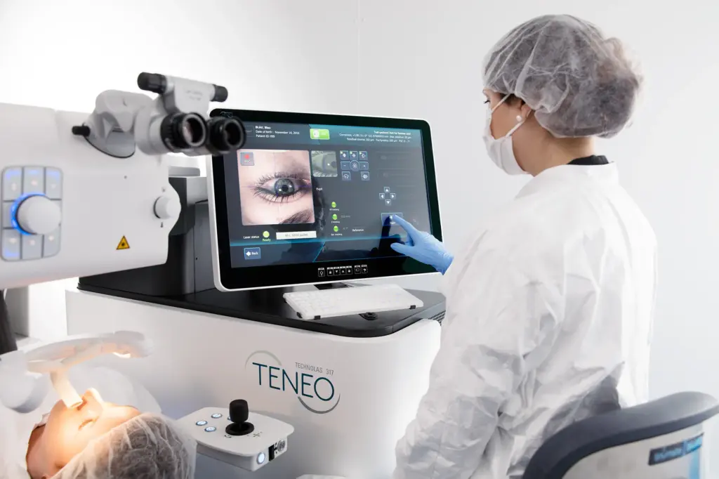 Medical professional in cleanroom attire operates a TECHNOLAS TENEO 317 laser eye surgery machine. A patient lies ready for treatment while their eye is displayed on the monitor.