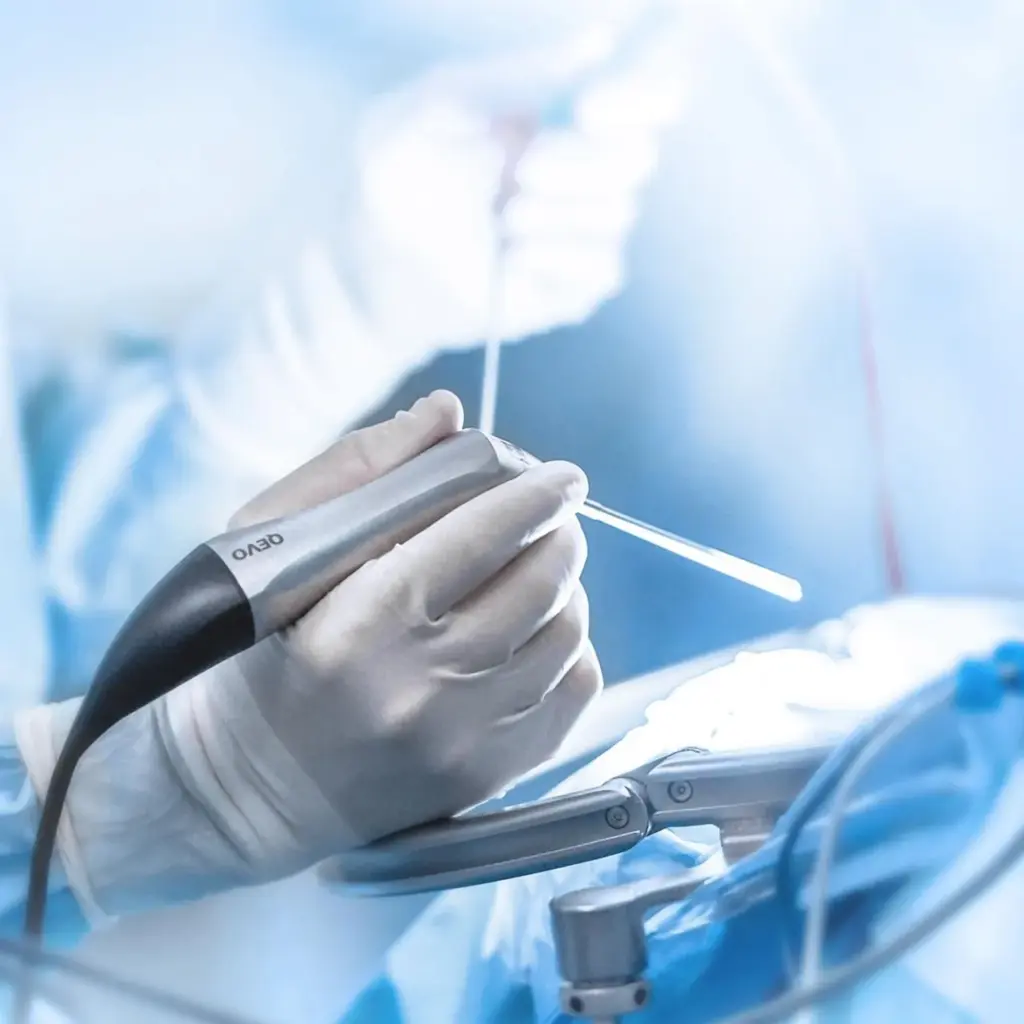 Gloved hand holding a surgical instrument, likely a drill, in an operating room with bright lights illuminating the sterile surgical field.