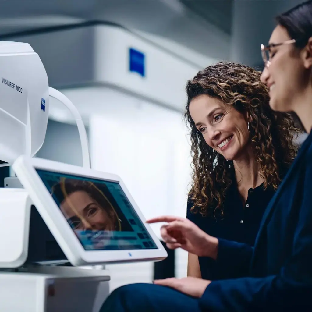 An optician assists a smiling patient, pointing at a ZEISS optical device screen showing a facial scan for precise measurements.
