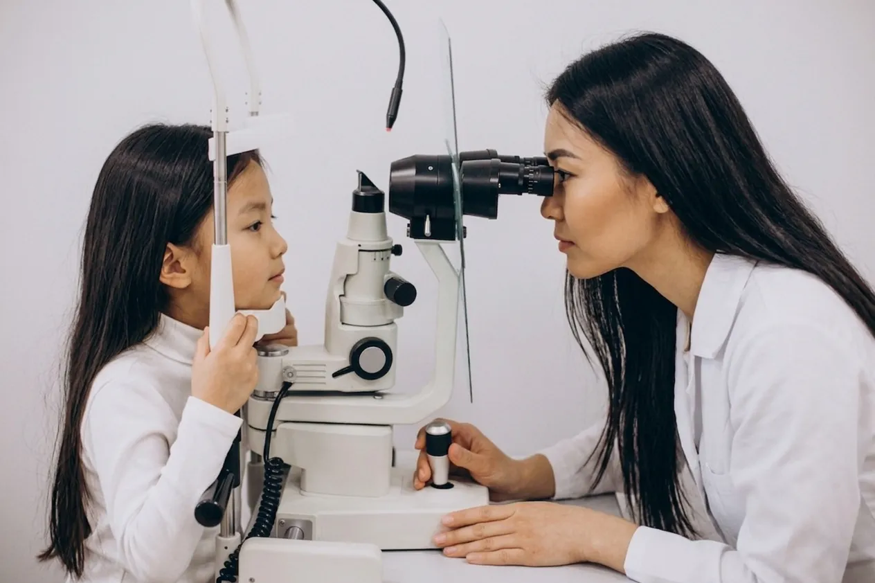 An Asian female optometrist in a white lab coat uses a slit lamp to examine the eye of a young Asian girl with her head supported.